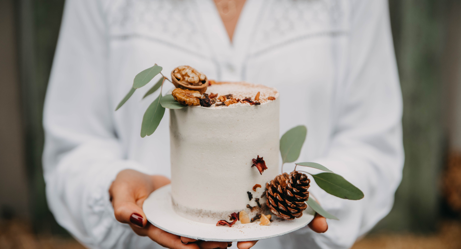 A person in a white tunic holds a small white cake decorated with autumn-themed elements. Another person is holding a small round white cake adorned with dried leaves and chestnuts.