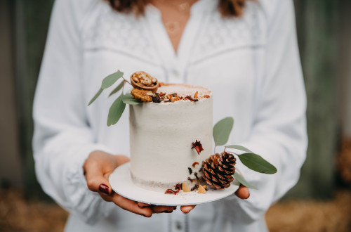 A person in a white tunic holds a small white cake decorated with autumn-themed elements. Another person is holding a small round white cake adorned with dried leaves and chestnuts.