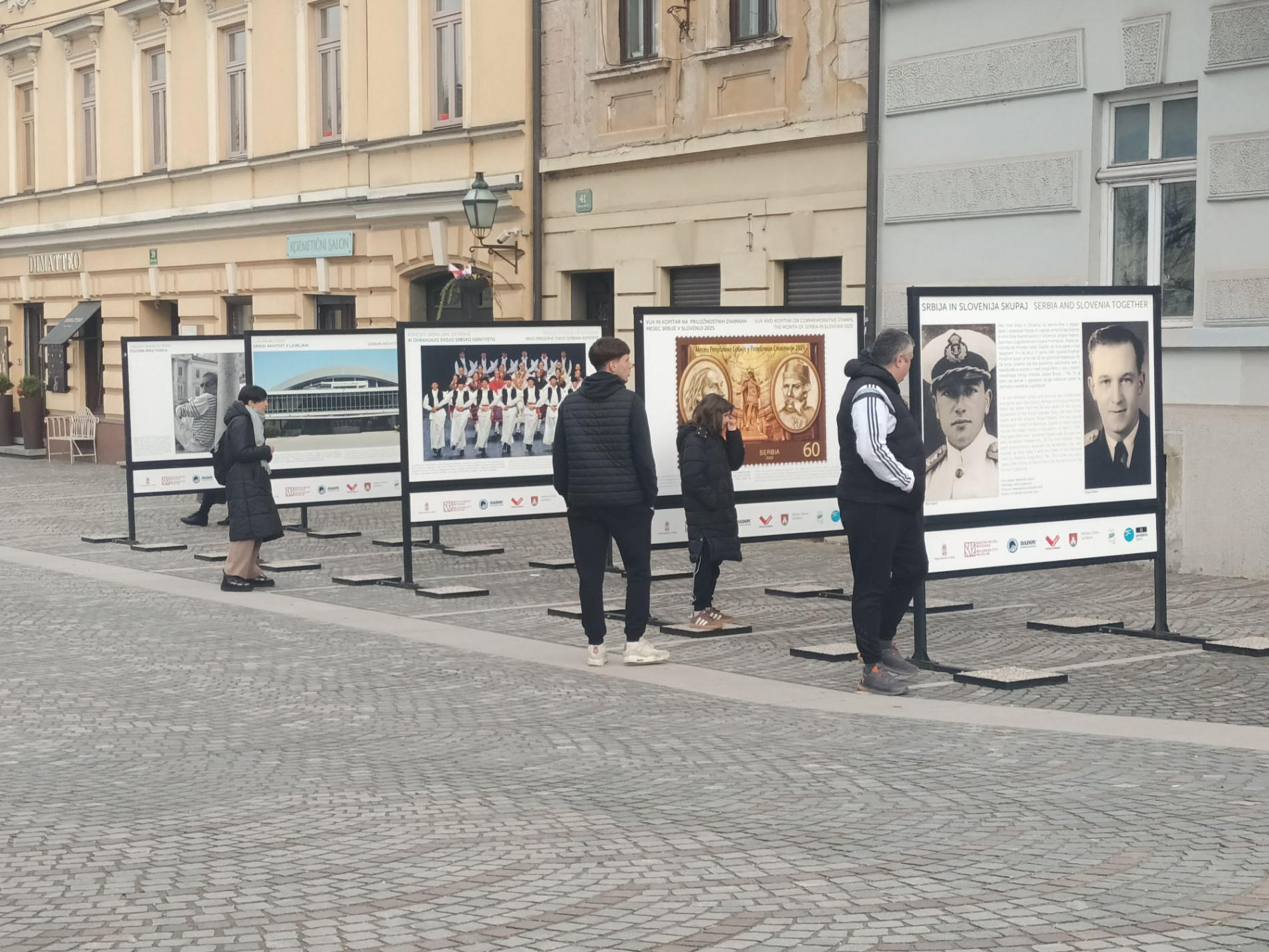 Obiskovalci si ogledujejo razstavne panoje na prostem na Gallusovem nabrezju med zgodovinskimi stavbami.