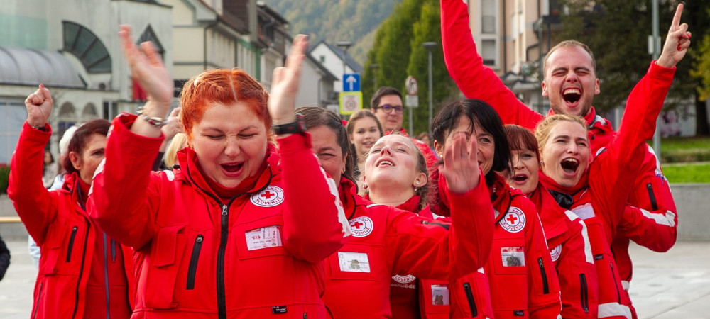 A group of Red Cross volunteers in red jackets enthusiastically cheer and clap on a street.