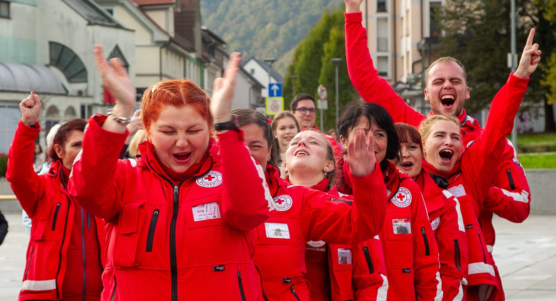 Skupina prostovoljcev Rdecega kriza v rdecih jaknah na ulici navduseno vzklika in ploska.