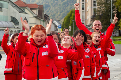 Skupina prostovoljcev Rdecega kriza v rdecih jaknah na ulici navduseno vzklika in ploska.