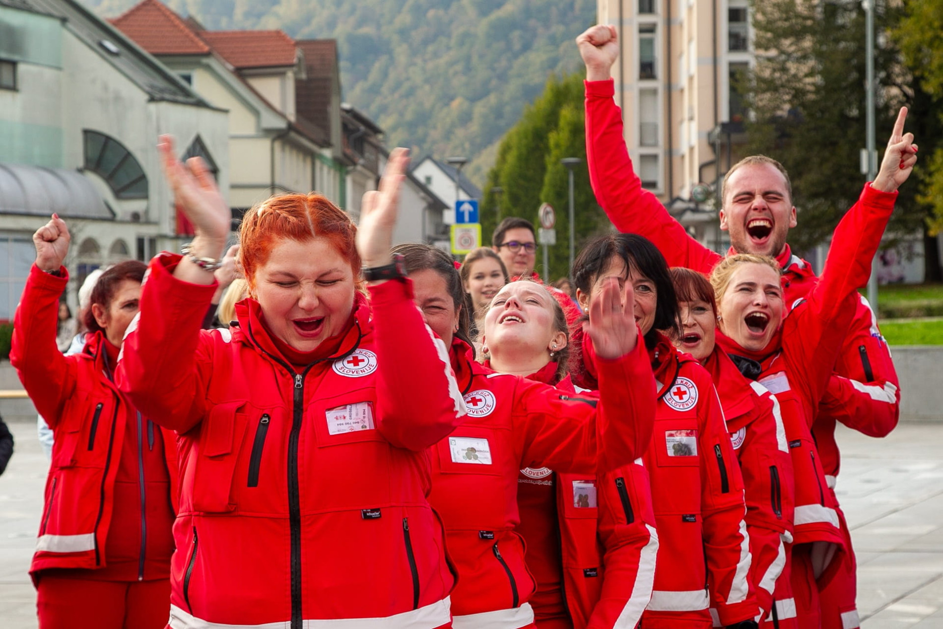 Skupina prostovoljcev Rdecega kriza v rdecih jaknah na ulici navduseno vzklika in ploska.
