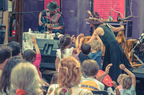 A couple with hats on their heads performs in front of an audience, where children are raising their hands in the air.