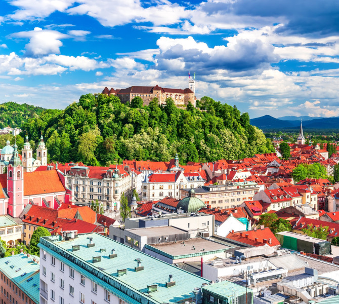 A view of rooftops of a city. Green hill with a castle on top reigning over the town. Blue sky.   