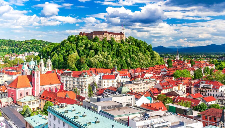 A view of rooftops of a city. Green hill with a castle on top reigning over the town. Blue sky.   
