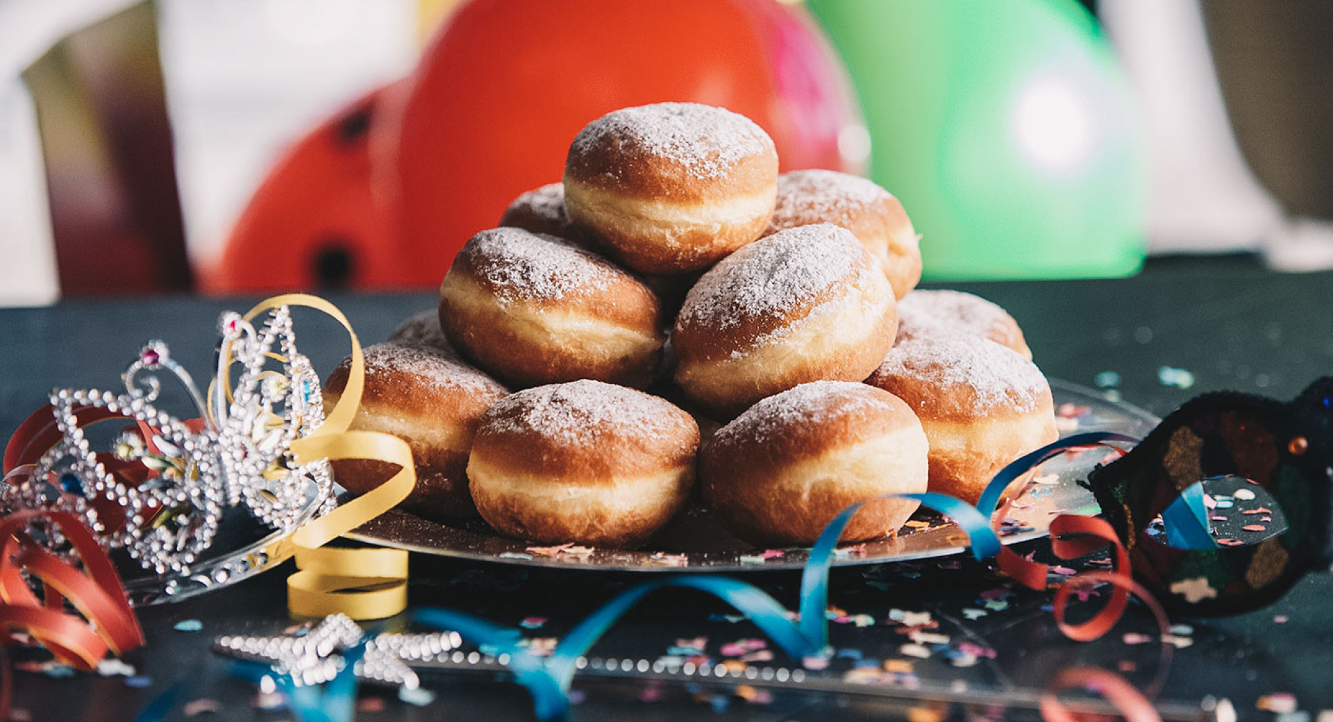 A large pile of donuts on a plate. The plate is on a black table decorated with confetti, colorful ribbons, and a crown. In the background, colorful balloons are out of focus.