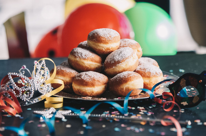 A large pile of donuts on a plate. The plate is on a black table decorated with confetti, colorful ribbons, and a crown. In the background, colorful balloons are out of focus.