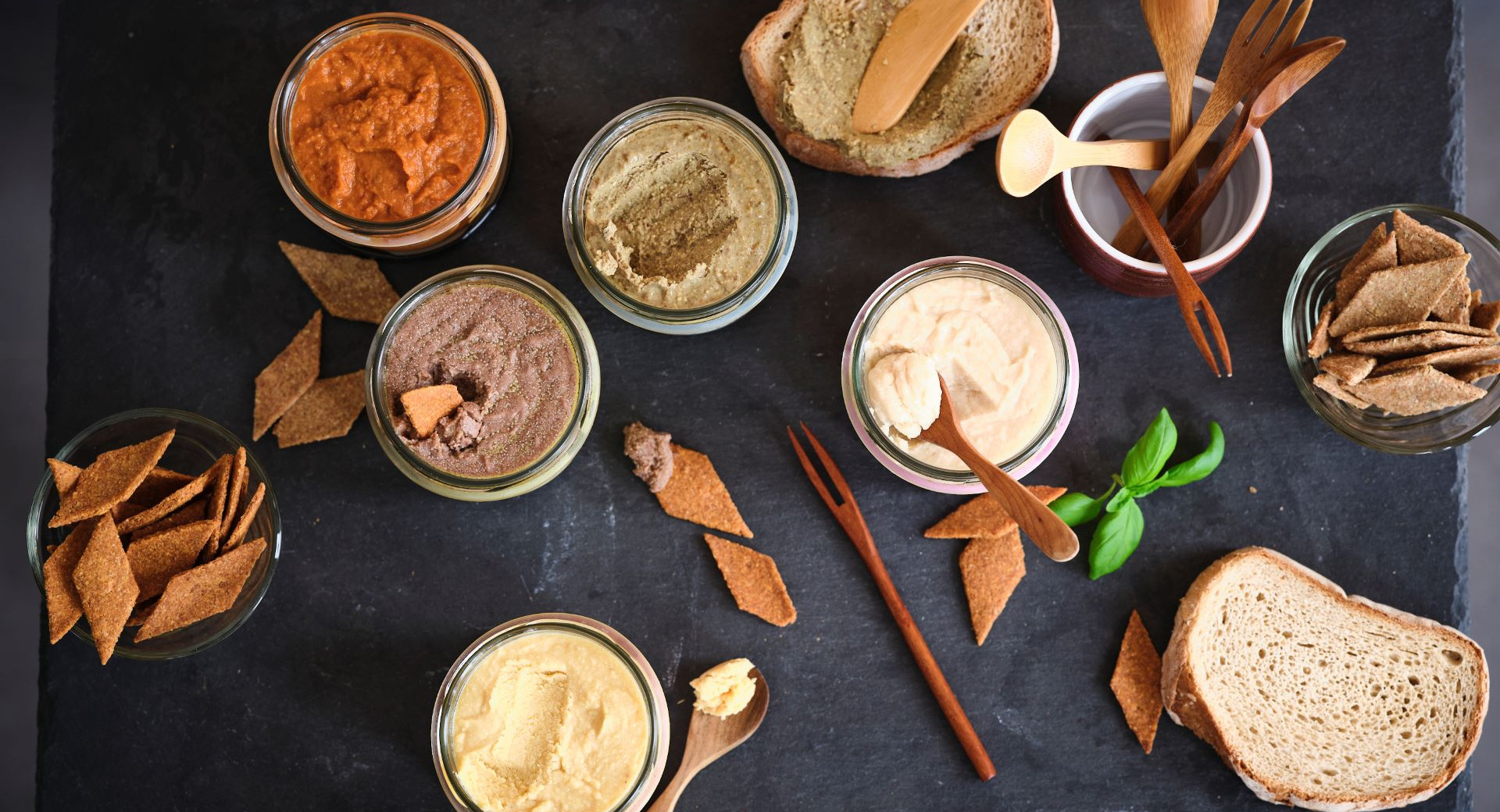A variety of plant-based spreads in jars, served with bread, crackers, and wooden cutlery on a black snack board.