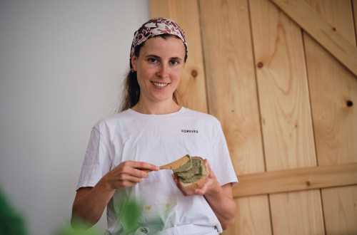 A smiling woman in a white T-shirt is spreading plant-based spread on bread in a cosy home setting with a wooden backdrop.