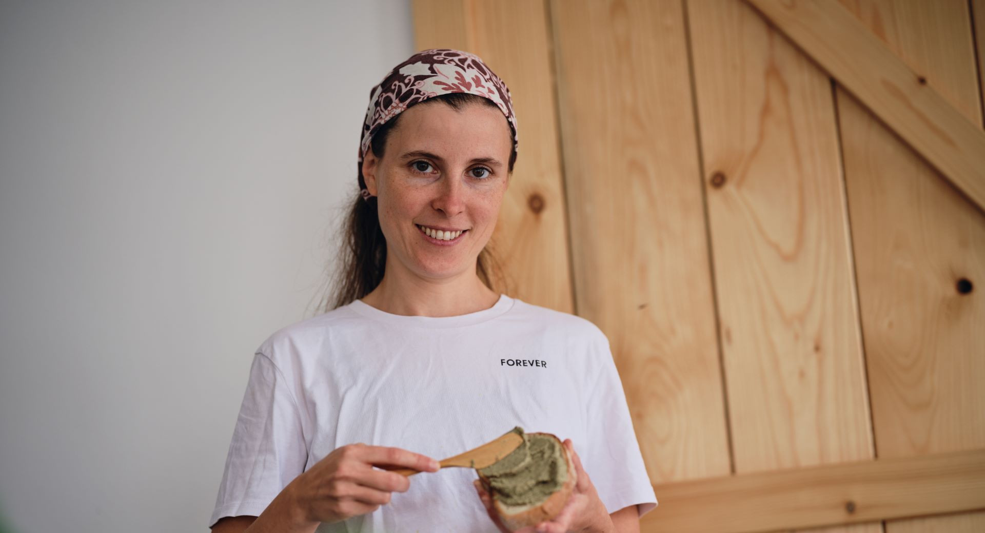 A smiling woman in a white T-shirt is spreading plant-based spread on bread in a cosy home setting with a wooden backdrop.