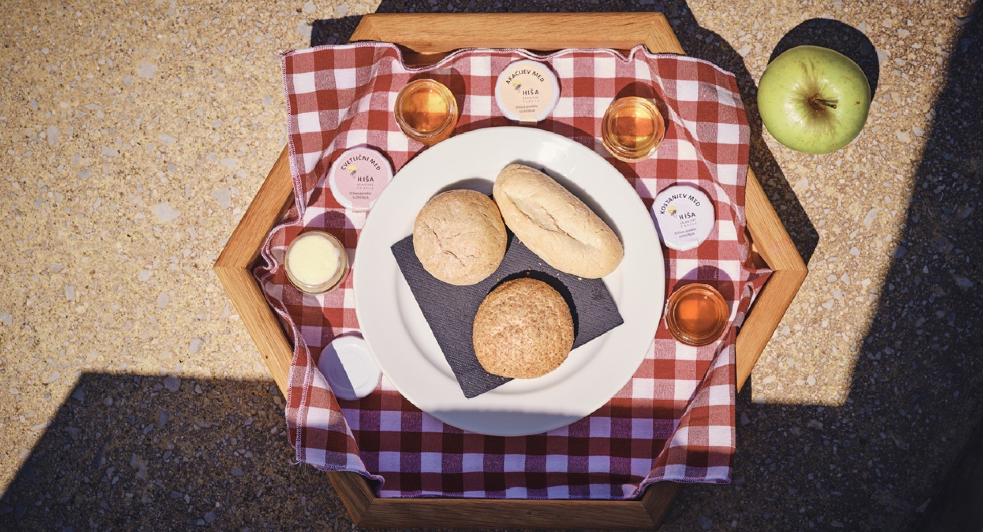 A checkered tablecloth with jars of honey, an apple, butter, and bread on a tray.