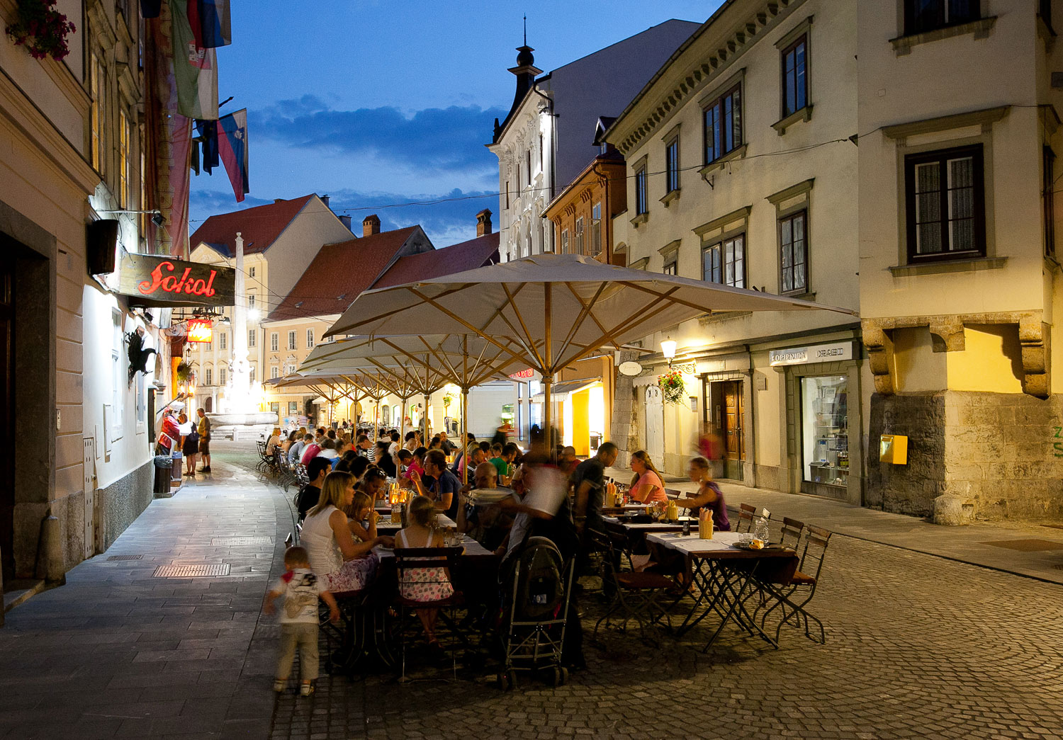 Gostje zvečer sedijo za mizami pod senčniki na terasi pred lokalom.