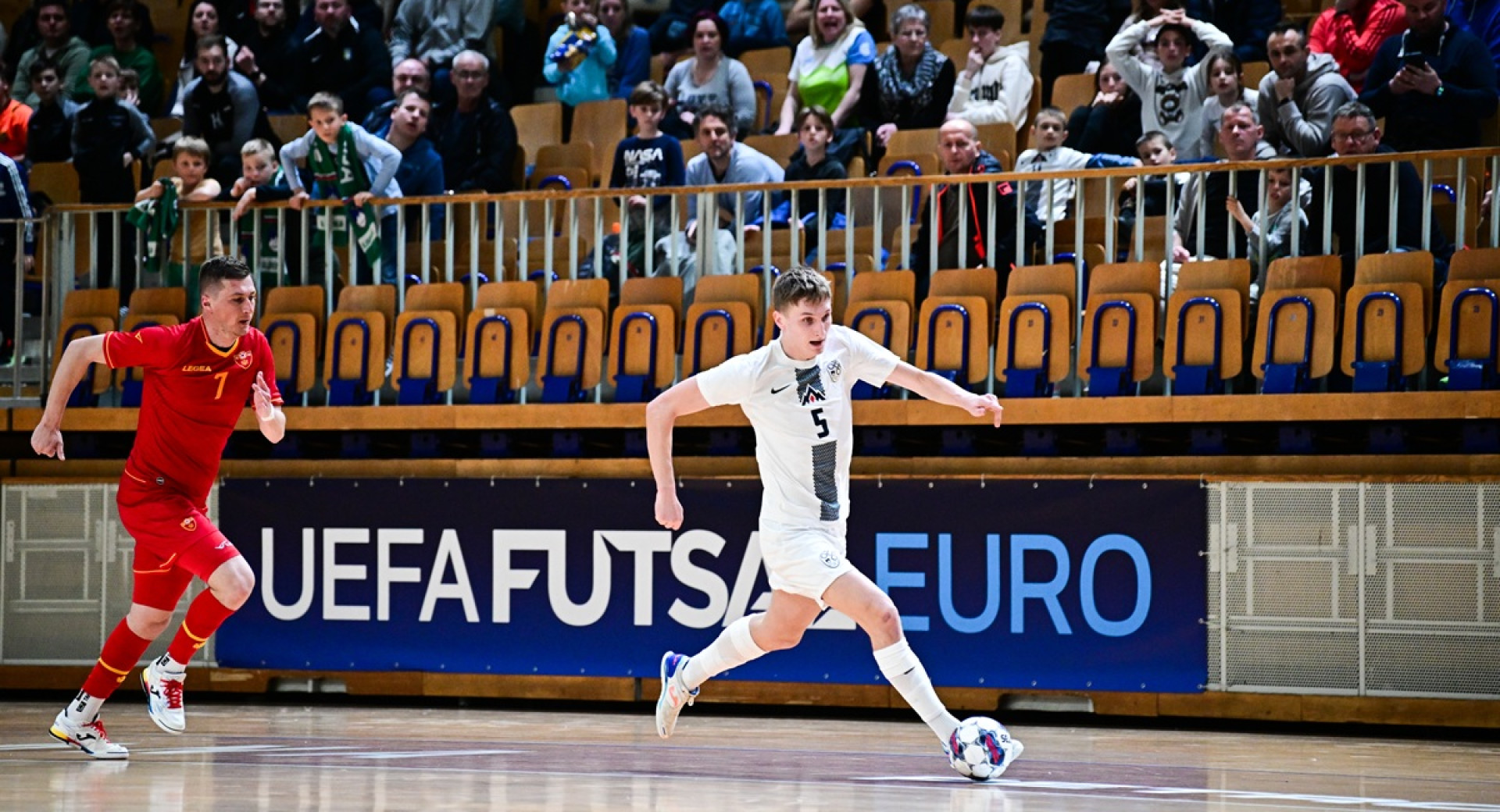Men in red and white jerseys running after the ball in front of a packed grandstand.