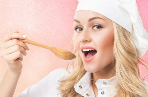 A woman with blue hair is wearing a white chef's outfit, holding a ladle in her right hand and bringing it towards her open mouth. She is posing in front of a pink backdrop.