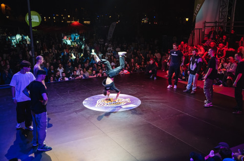 Fant izvaja breakdance in se vrti na rokah v sredini kroga, okoli njega pa so ostali plesalci, ki jih opazuje gruca gledalcev.