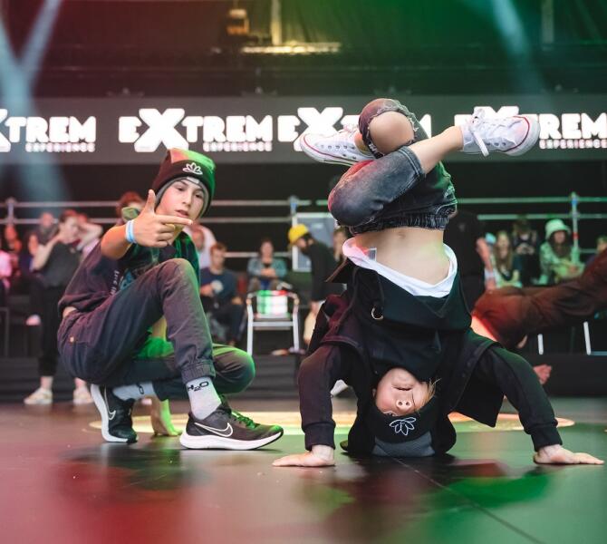 Two boys are breakdancing on stage – one is spinning on his head, while the other, crouching, points at him.