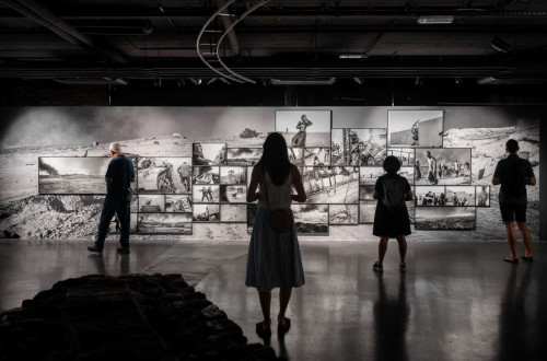 Osvetljena crno-bela fotografska razstava v temi; stirje opazovalci v senci si ogledujejo kolaz teh fotografij na steni.