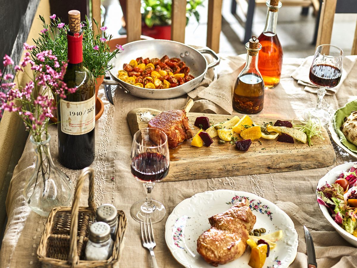 A rustic table set with wine, roasted meat, fresh vegetables, and herbs.