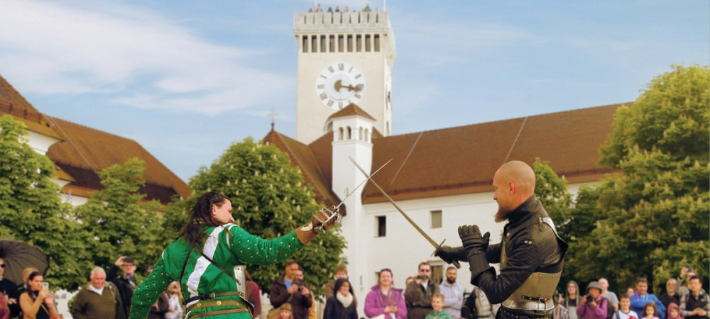 Two men in knightly attire are fighting with swords, with a view of the Observation Tower at Ljubljana Castle behind them.