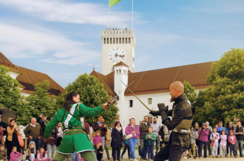 Dva moza v viteskih oblekah se bojujeta z meci, za njima pa je pogled na Razgledni stolp na Ljubljanskem gradu.
