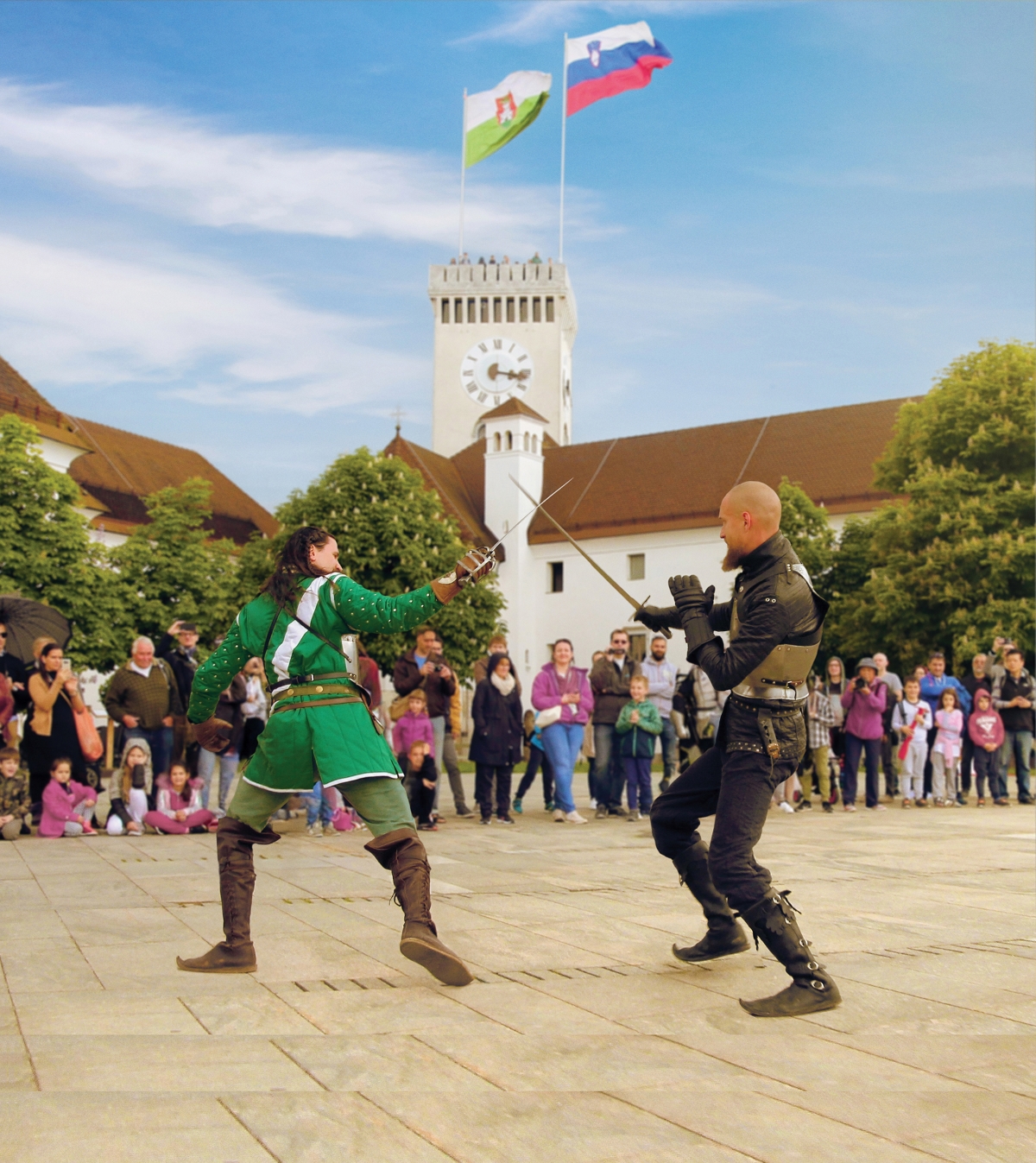Two men in knightly attire are fighting with swords, with a view of the Outlook Tower at Ljubljana Castle behind them.
