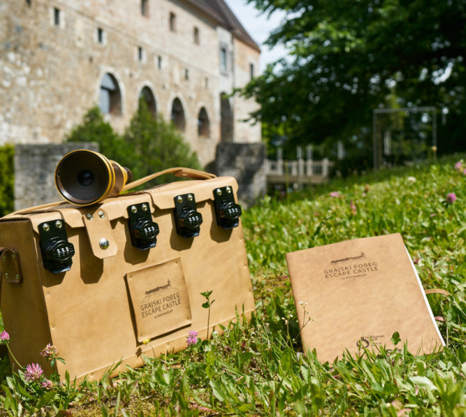 Mapa in kovcek s kljucavnico s kodo v bez barvi na travi, v ozadju pa Ljubljanski grad.