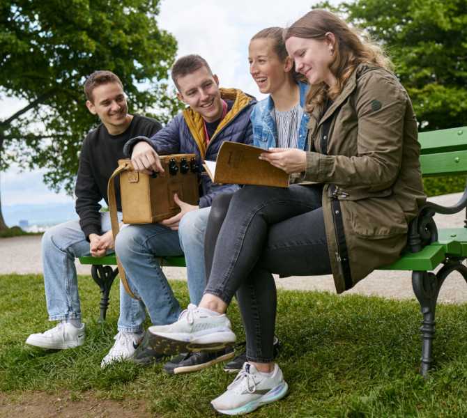 Skupina stirih ljudi sedi na zeleni klopci na zeleni travi in pregleduje mapo.