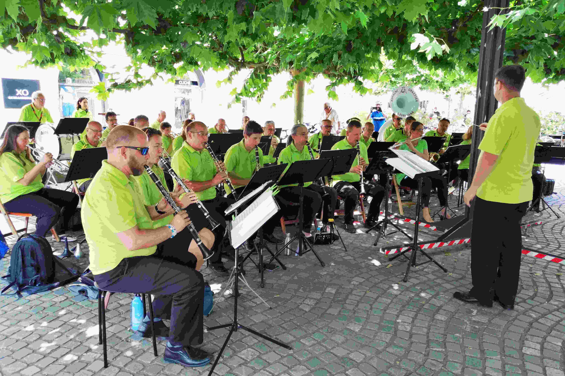 Pihalni orkester v fluorescentno zelenih majicah sedi pod drevesi in igra na glasbila.