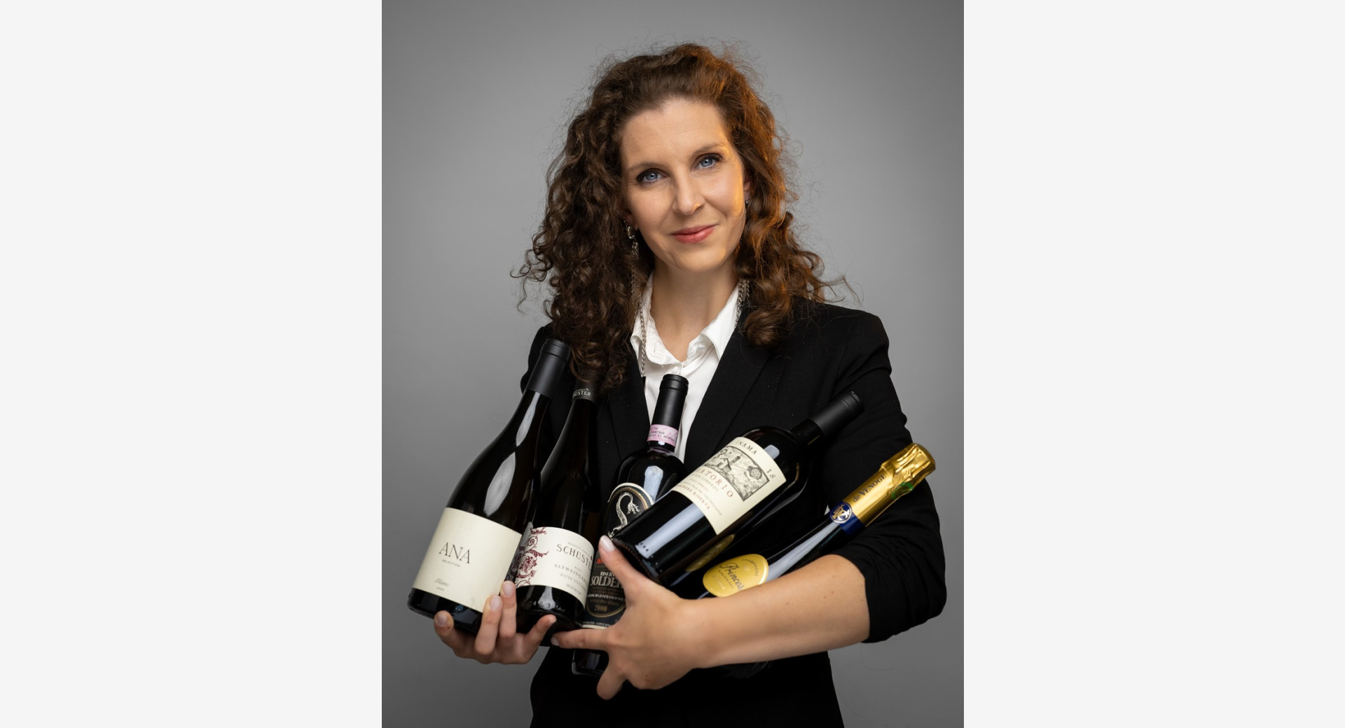 A woman with curly hair is holding five bottles of wine in her arms.