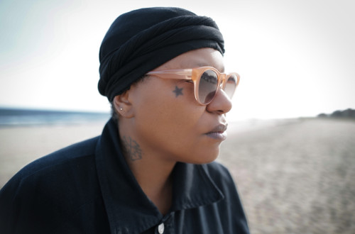 Close-up of a woman’s right profile, wearing a black shirt and black cap, with sunglasses and a star tattoo beside her right eye; in the background, a blurred sea and sandy beach.