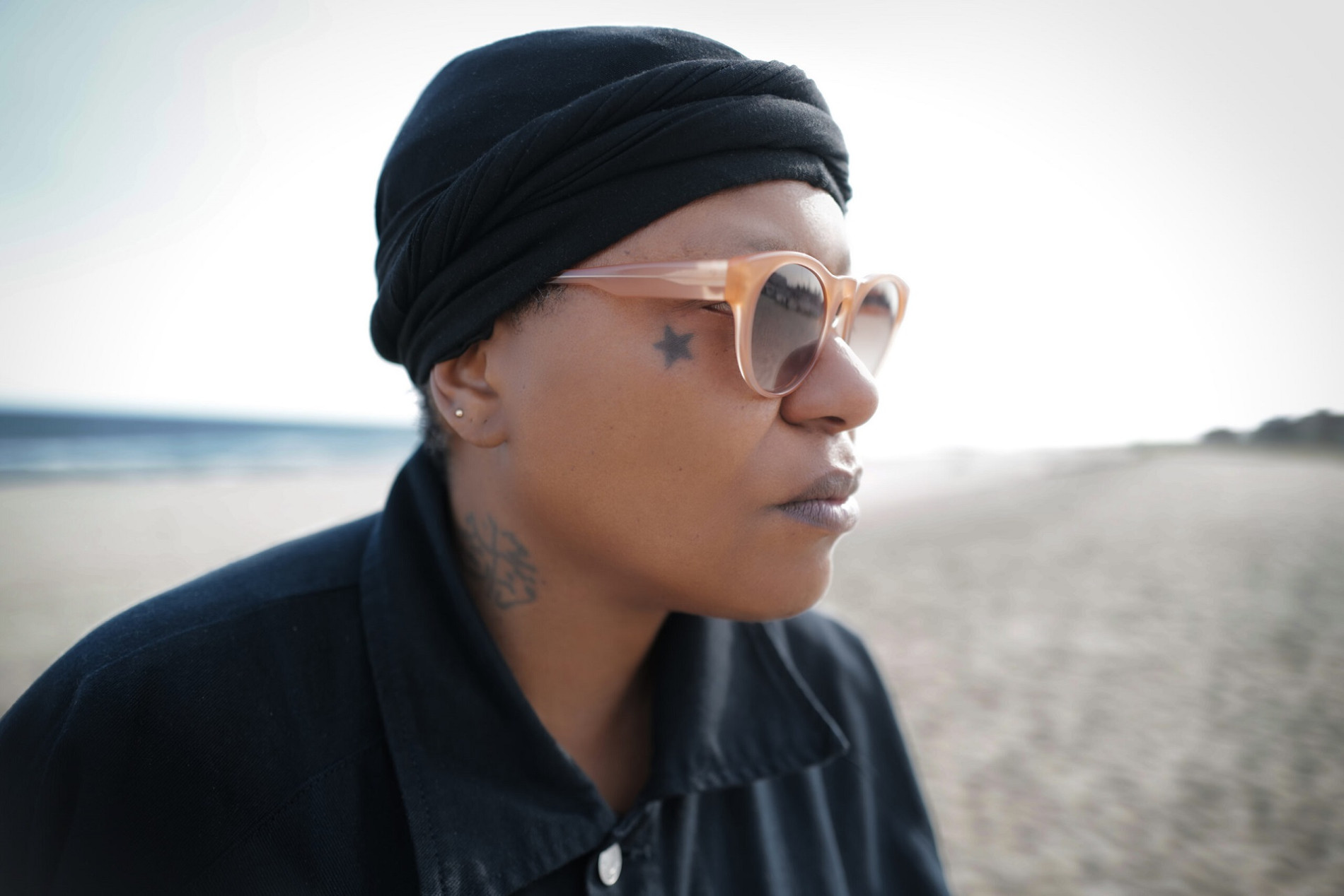 Close-up of a woman’s right profile, wearing a black shirt and black cap, with sunglasses and a star tattoo beside her right eye; in the background, a blurred sea and sandy beach.