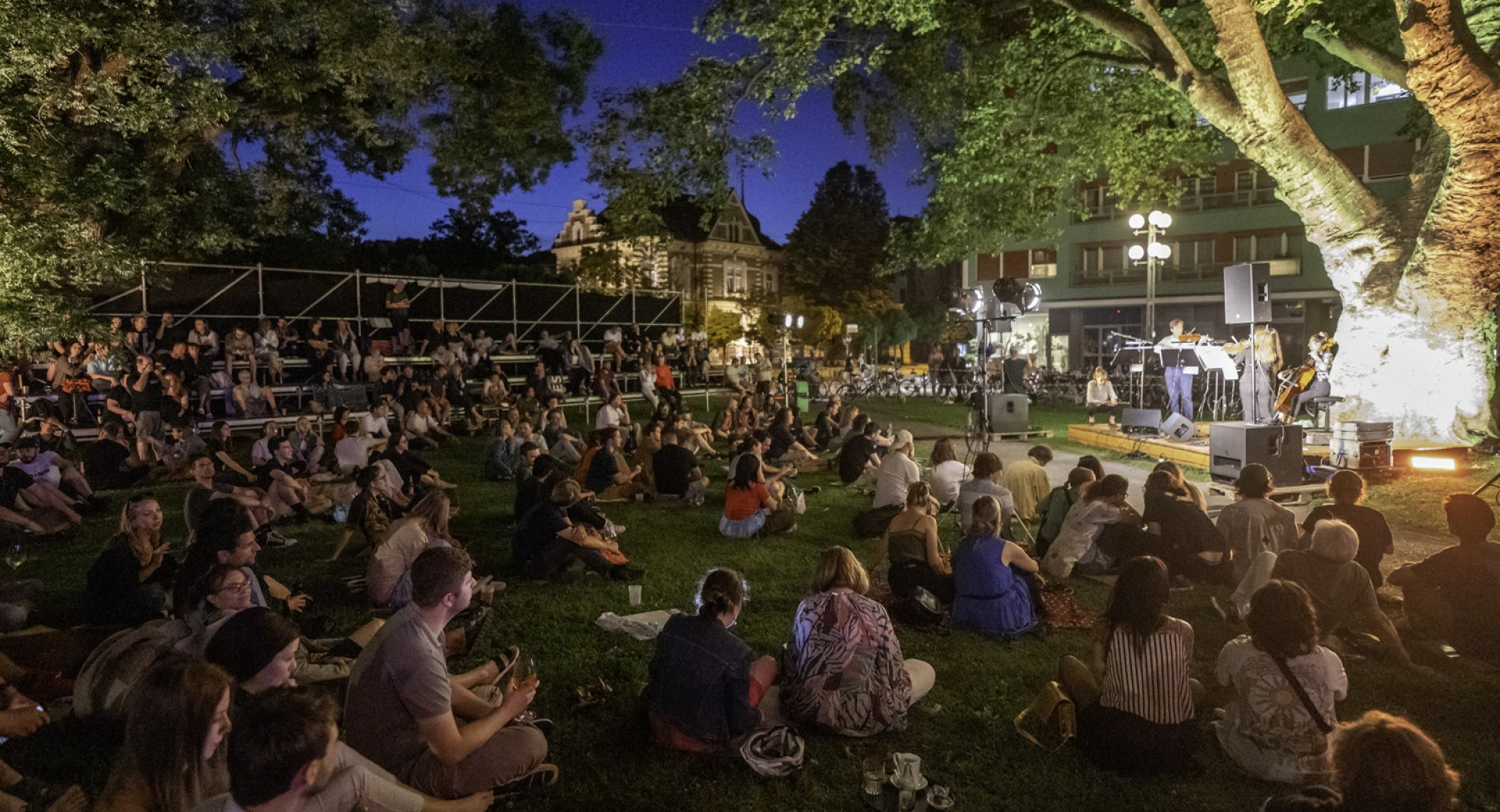 At dusk, people are sitting on the meadow, watching and listening to a concert.