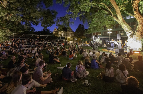 At dusk, people are sitting on the meadow, watching and listening to a concert.