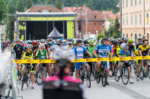 A group of cyclists is preparing for the start of a race, surrounded by the energetic atmosphere of the event and urban architecture in the background.