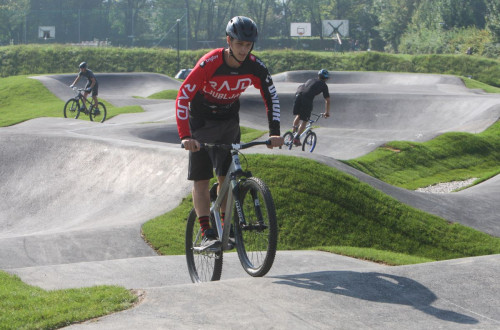 A young man on a bike riding on a pump track, surrounded by green hills, with other cyclists visible in the background navigating the undulating paths.