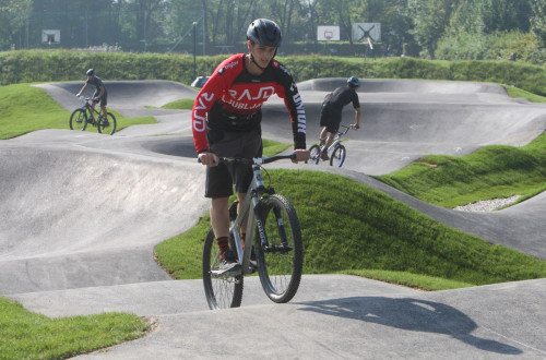 A young man on a bike riding on a pump track, surrounded by green hills, with other cyclists visible in the background navigating the undulating paths.