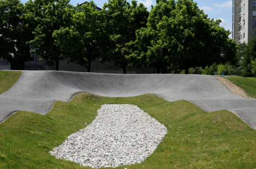 A pump track with characteristic undulating paths, surrounded by green grass and sprinkled with white pebbles, set against a backdrop of trees.