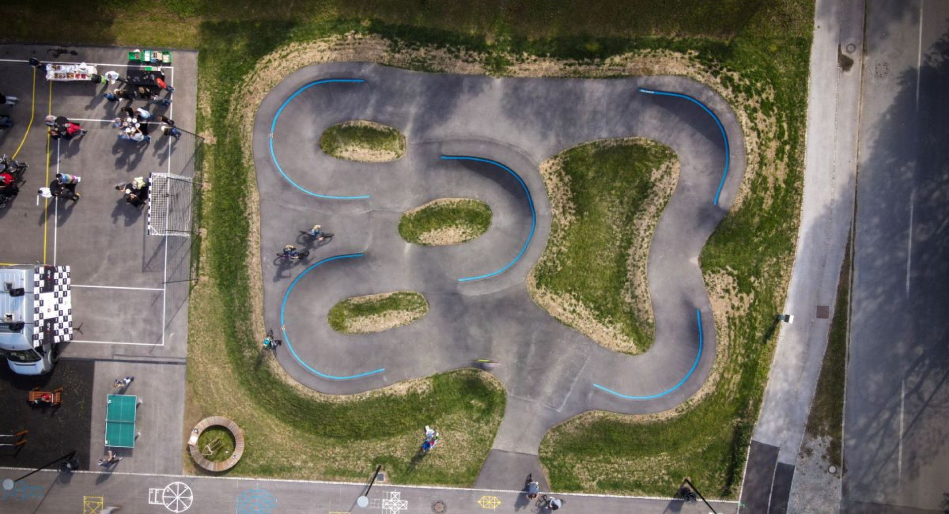 An aerial view of a pump track with flowing curves and blue marking lines, surrounded by green grass and adjacent spectator areas.