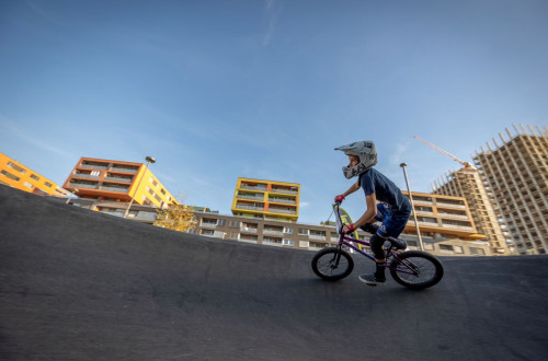 The photo features a young cyclist with a BMX on a pump track, surrounded by modern, colorful apartment buildings and a construction crane in the background.
