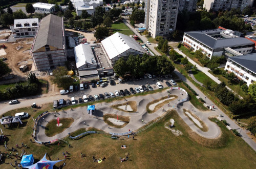 An overhead view of the pumptrack track with surrounding buildings, cars and an event where visitors gather under the floors.
