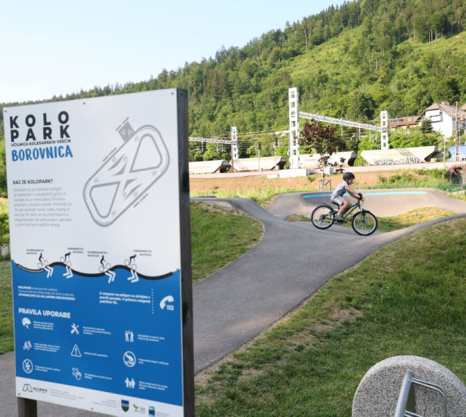 A cyclist on a pump track with an informational sign reading "Kolopark Borovnica," surrounded by nature and nearby infrastructure.
