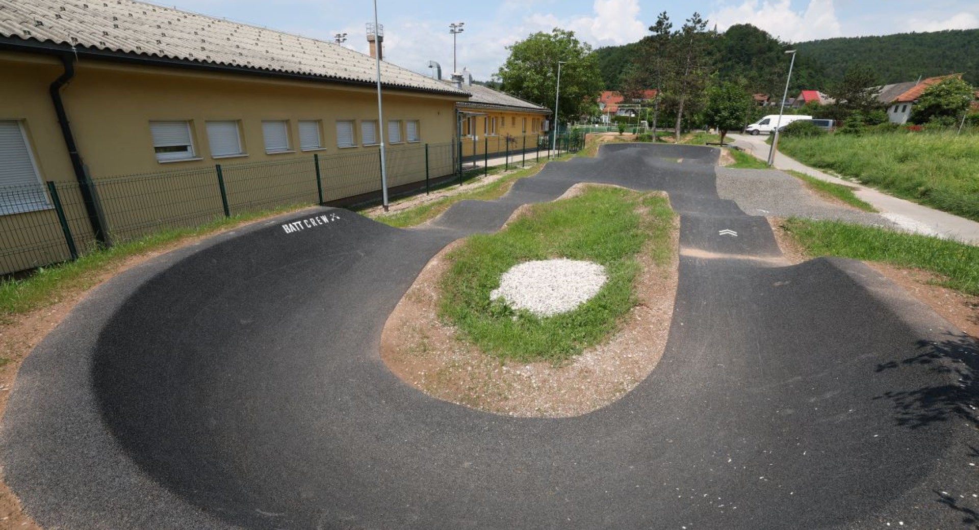 Pumptrack track next to the building in sunny weather, surrounded by greenery and a varied surface for cycling.