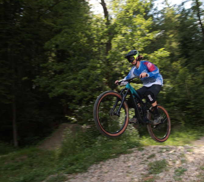 A cyclist performs a jump on a mountain bike in a forest surrounded by trees.