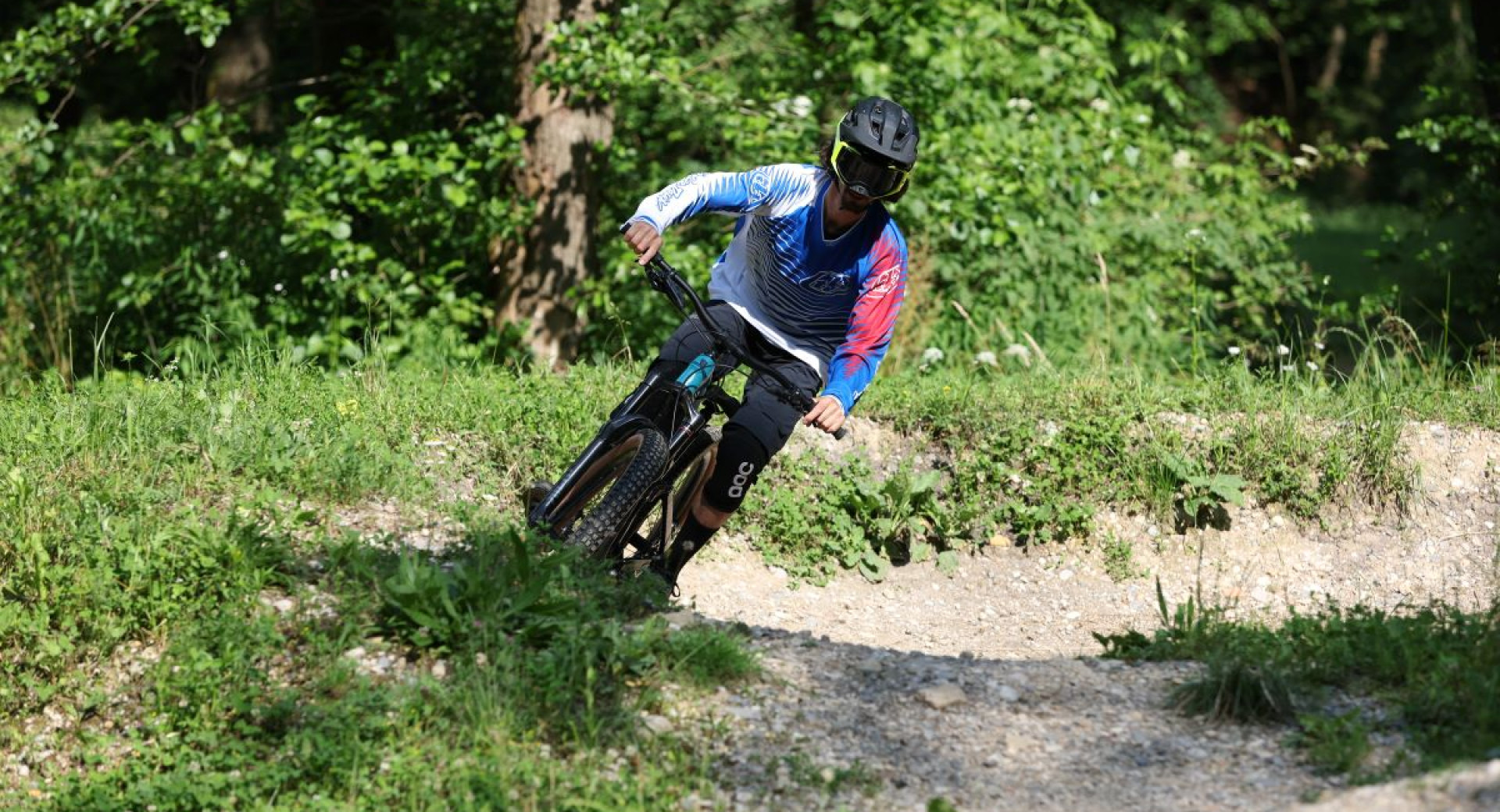 A cyclist on a forest path between turns, surrounded by greenery and trees.