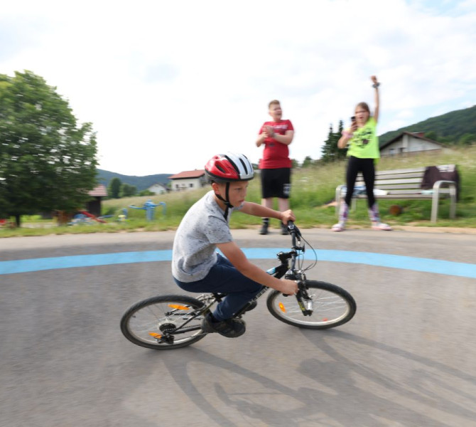 A boy on a bicycle rides on a paved cycling track, while people in the background watch and encourage him.