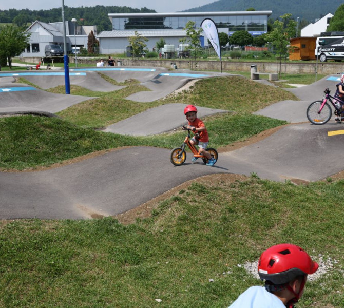 A child rides on a pushchair on the undulating terrain of a pumptrack, surrounded by nature and cycling equipment.
