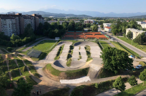An urban environment with a BMX track, surrounded by green surfaces and residential buildings, with views of distant mountains in the background.