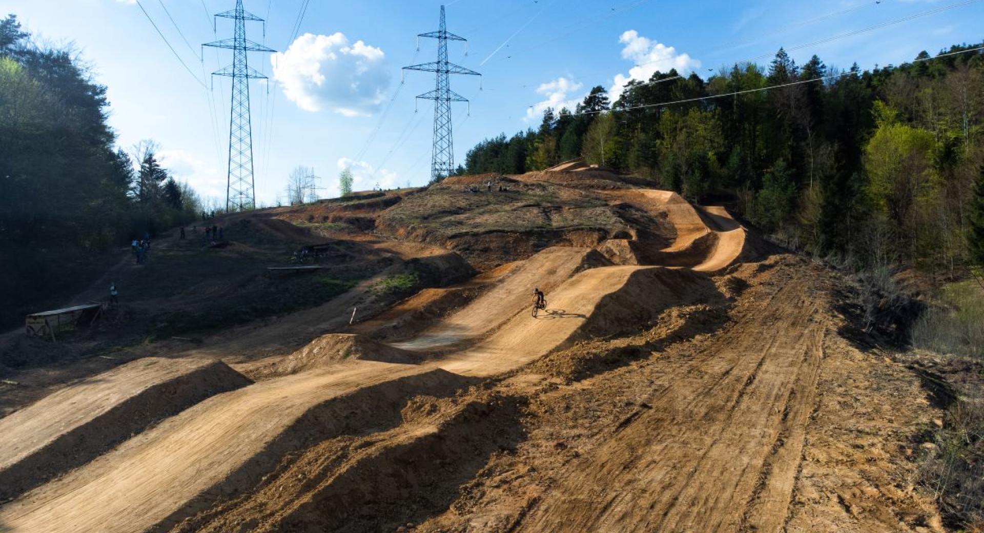 A mountain bike course with undulating terrain, surrounded by forests, under power lines and on a sunny day.