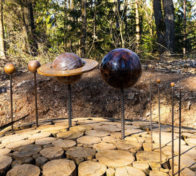 Kovinska umetniska upodobitev planetov soncnega sistema, vkljucno s Saturnom z obrocem, postavljena na lesenih okroglih ploscah v gozdu.